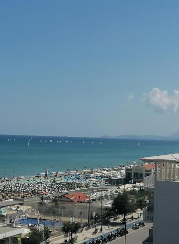 Spiaggia affollata con ombrelloni, mare calmo e barche a vela all'orizzonte.