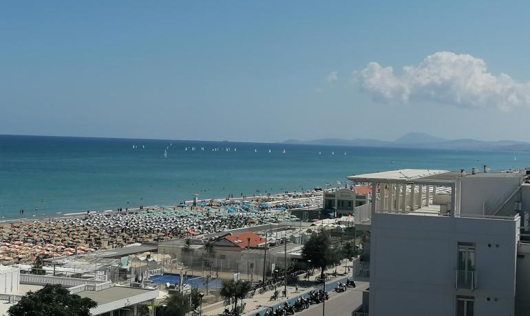 Spiaggia affollata con ombrelloni, mare calmo e barche a vela all'orizzonte.