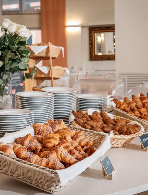 Colazione a buffet con croissant, torta e piatti, decorata con fiori bianchi.