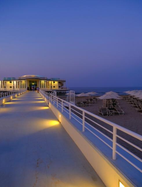 Lungomare al tramonto con passerella illuminata e ombrelloni sulla spiaggia.