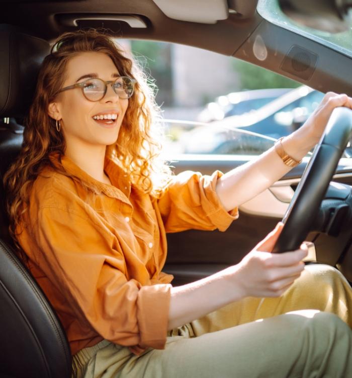 Donna sorridente con occhiali guida un'auto, indossa camicia arancione e orologio.