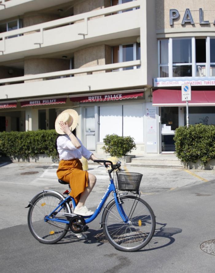 Donna in bicicletta davanti all'Hotel Palace, indossa cappello e gonna arancione.