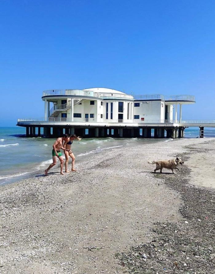 Due persone e un cane sulla spiaggia, vicino a un edificio sul mare.