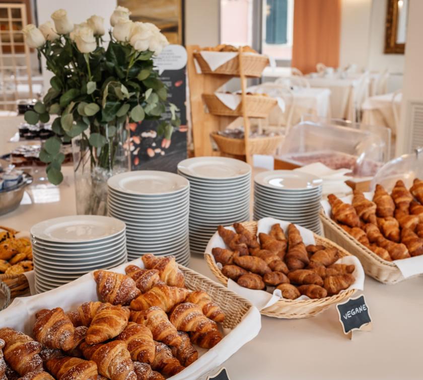 Buffet colazione con croissant, piatti impilati e rose bianche in un vaso.