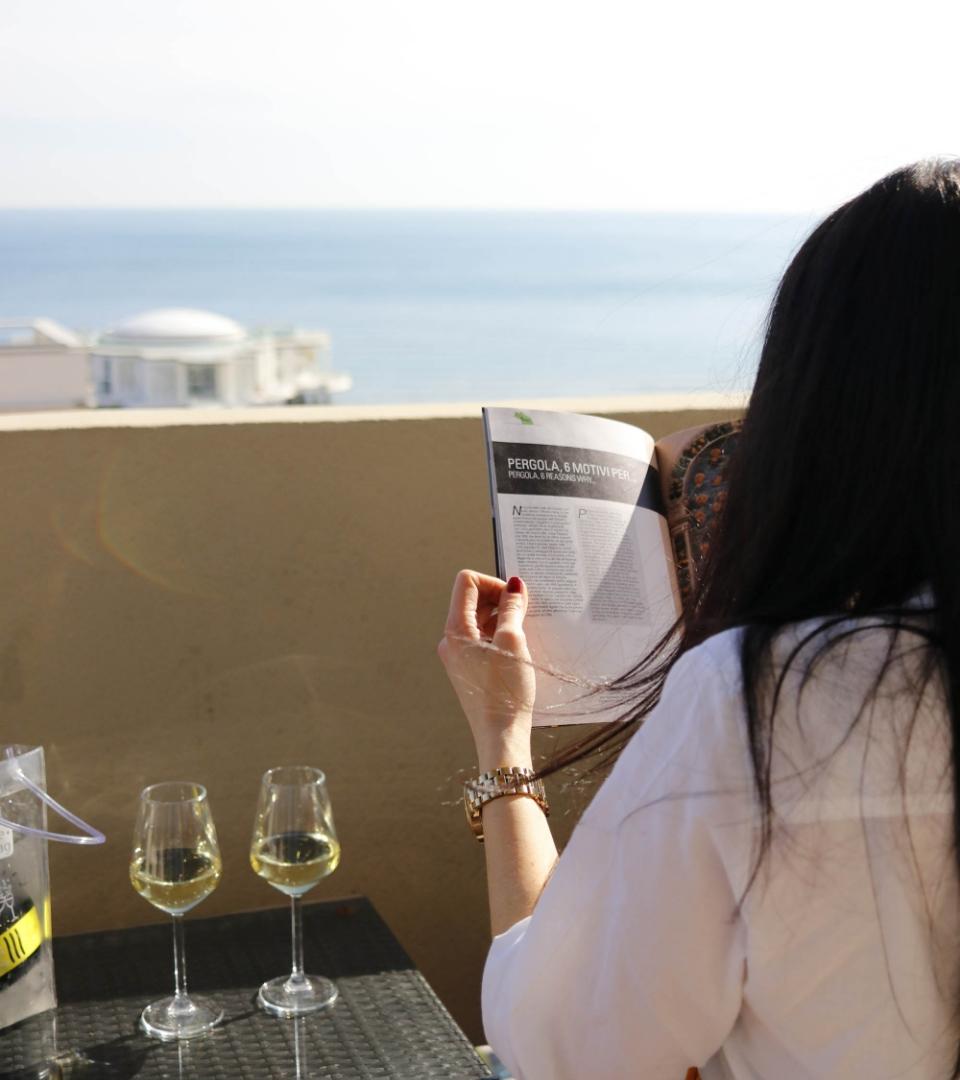 Donna legge rivista su terrazza con vista mare, due calici di vino bianco.