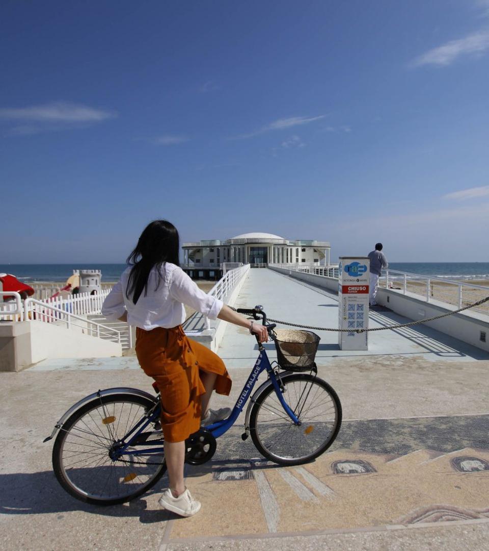 Donna in bicicletta guarda il mare e un edificio bianco su un molo.