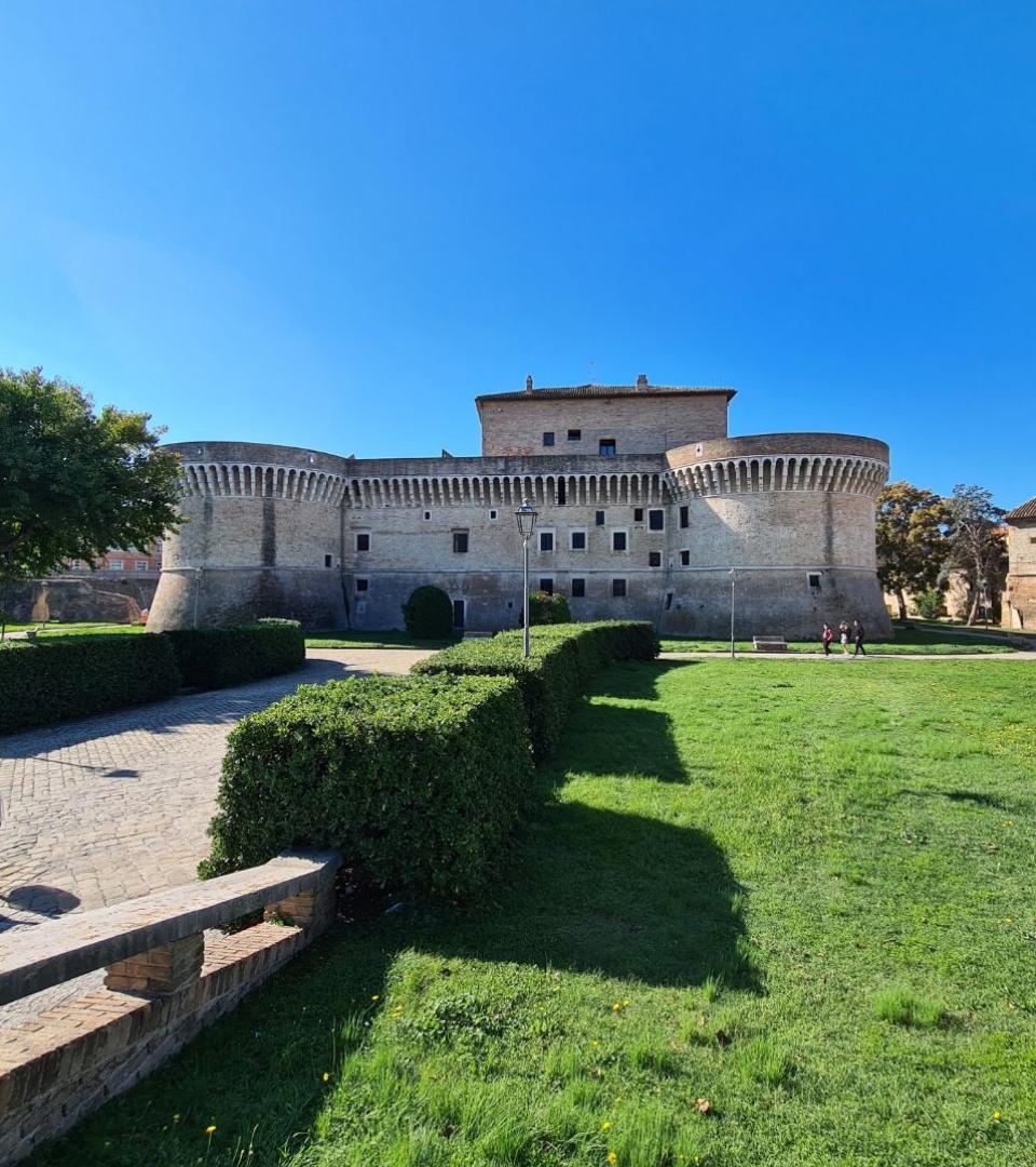 Antica fortezza con torri circolari, circondata da un giardino verde e cielo limpido.