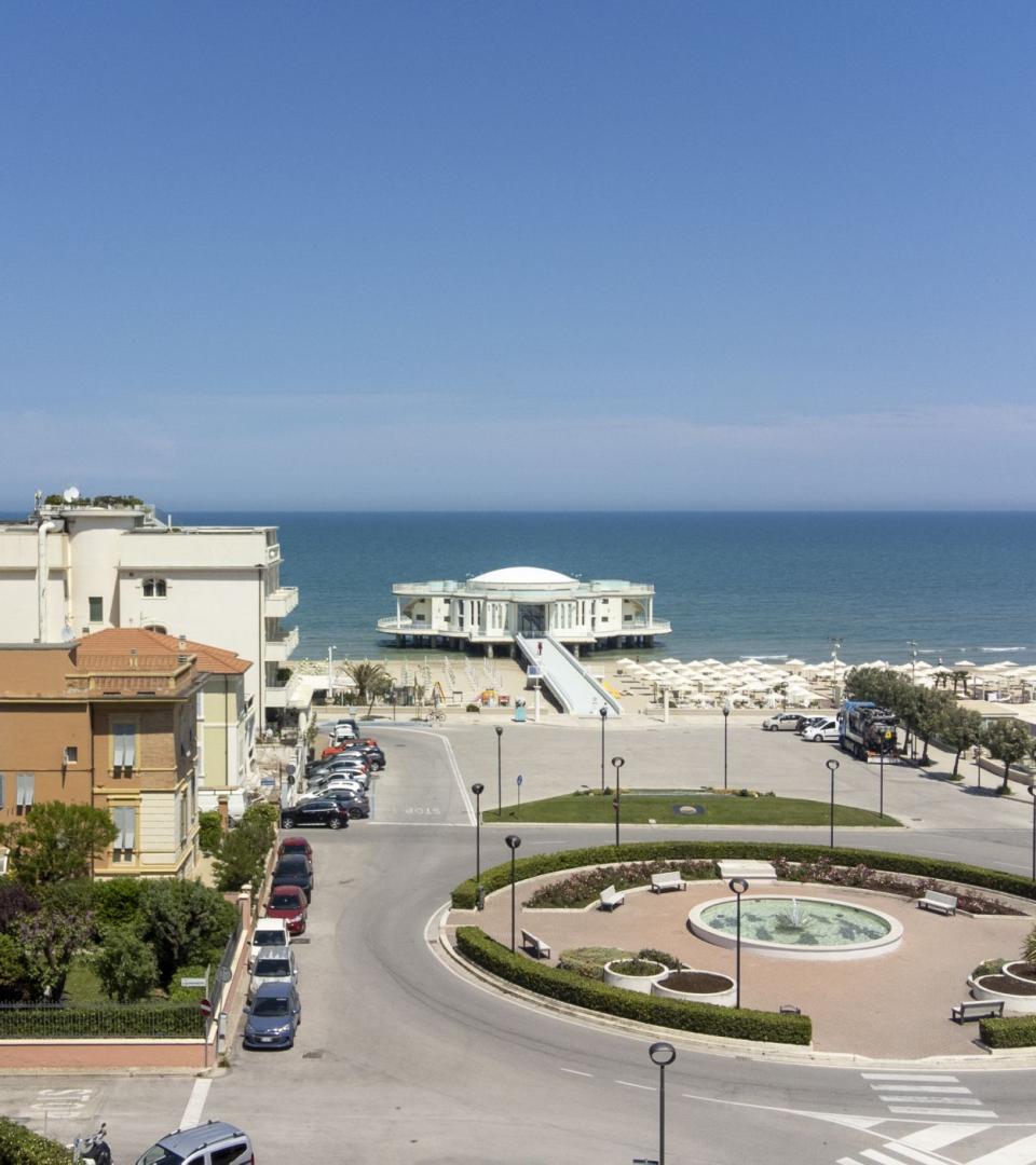 Vista sul mare con edificio circolare e rotonda, spiaggia attrezzata sullo sfondo.